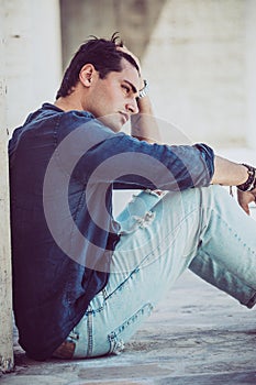 Young man sitting on the ground outdoors with hand in hair. Casual clothes, profile view
