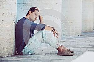 Young man sitting on the ground outdoors with hand in hair. Casual clothes, profile view