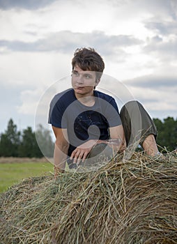 A young man is sitting on a haystack against a cloudy sky.