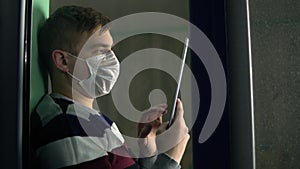 A young man is sitting at home in quarantine with a tablet in his hands. A man sits on a window sill in a mask and works