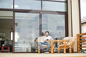 Young man sitting on house terrace chair, relaxing using phone