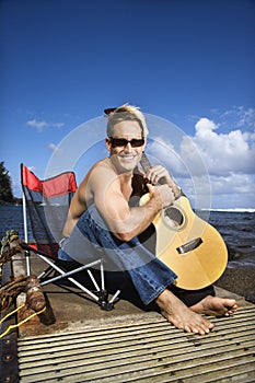 Young Man Sitting Lakeside and Holding Guitar