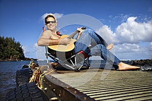 Young Man Sitting Lakeside and Playing Guitar