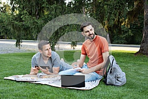 young man is sitting with a laptop on grass on a college campus and taking notes in notebook. Man explaining something to her fri photo