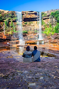 young man sitting near waterfall falling from mountain top with reflection and blue sky