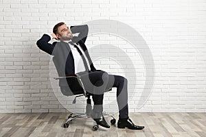 Young man sitting in office chair near white brick wall indoors, space for text