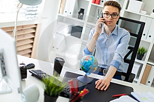 A young man is sitting in the office at a computer desk and talking on the phone. Before the young man on the table is a