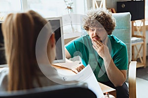 Young man sitting in office during the job interview with female employee, boss or HR-manager, talking, thinking, looks