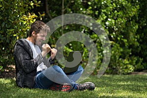 Young man sitting on a park while watching his computer and celebrating furiously with his hands. Natural Environment.