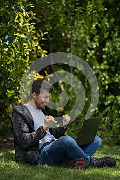 Young man sitting on a park while watching his computer and celebrating furiously with his hands. Natural Environment. Vertical