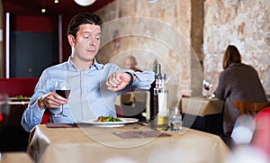 Young man sitting in restaurant with wine glass, looking at clock in anticipation of friend