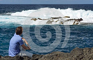 Young man sitting on rock shore and looking at ocean. View from behind. Blue ocean with high waves, water splash