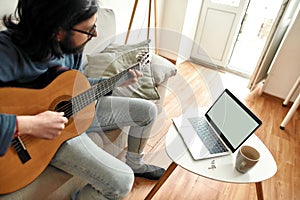 Young man sitting on sofa at home and learning how to play guitar, watching online course on laptop