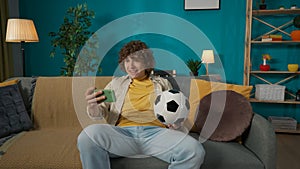 Young man sitting on the sofa in the room with a bootball ball and holding a smartphone in hand