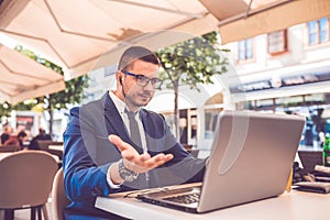 Young man sitting at street cafe having a conversation on a laptop wearing a headphones talking looks angry