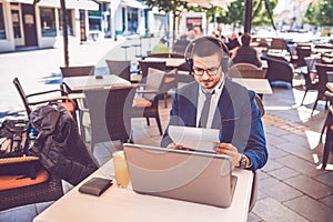 Young man sitting at  street cafe reading documents while working on laptop and wearing headphones