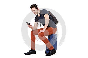 Young man sitting on suitcase and looking at mobile phone while listening to music