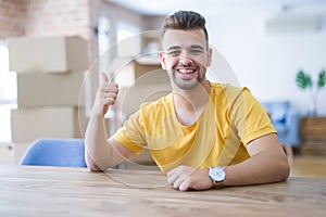 Young man sitting on the table with cardboard boxes behind him moving to new home smiling with happy face looking and pointing to