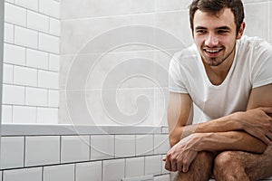 A young man is sitting on the toilet in the bathroom. He smiles
