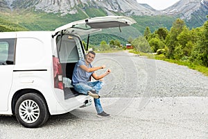 Young man sitting in the trunk of the car in the mountains background is relaxing and enjoy at a roadside. Car trip vocation. Car