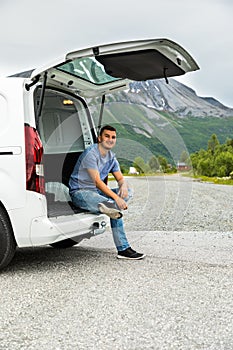 Young man sitting in the trunk of the car in the mountains background is relaxing and enjoy at a roadside. Car trip vocation. Car