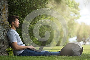 Young man sitting under a tree with his legs stretched and his computer on them. Technological concept.