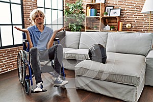 Young man sitting on wheelchair at for motorcycle accident clueless and confused expression with arms and hands raised