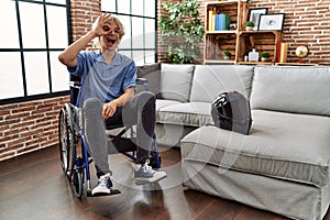 Young man sitting on wheelchair at for motorcycle accident smiling happy doing ok sign with hand on eye looking through fingers