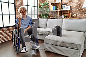 Young man sitting on wheelchair at for motorcycle accident smiling with happy face looking and pointing to the side with thumb up