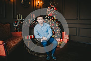 Young man sitting on the wooden horse near Christmas tree with presents and lights in dark living room