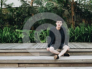 Young man sitting on wooden stair near the pool at holet yard with exotic plants at sunny day