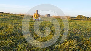 Young man sitting in yoga pose at green grass in the meadow and meditates. Muscular guy relaxing in lotus pose at nature