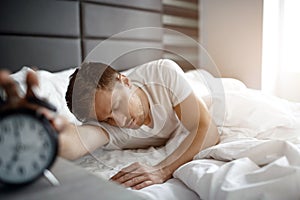 Young man sleep on bed in morning. He hold hand on clock. Oversleep. Daylight. photo