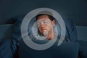 Young man sleeping in front of a laptop in a bed at home, tired after work day. Male fall asleep while watching movie on