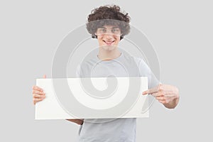 Young man, smiling and handsome, pointing with finger to show a blank white signboard, isolated on gray background. Placard copy