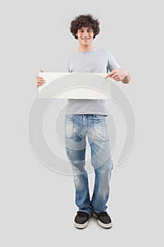 Young man, smiling and handsome, pointing with finger to show a blank white signboard, isolated on gray background. Placard copy