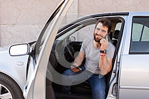 young man smiling and making a call on his phone, sitting on driver's seat with opened door