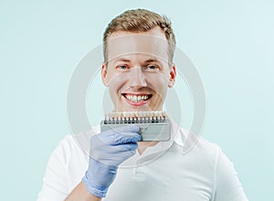 Young man smiling with natural white teeth in light blue background in dental clinic. Hands holding the teeth color palette next