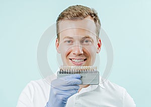 Young man smiling with natural white teeth in light blue background in dental clinic. Hands holding the teeth color palette next