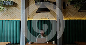 Young man smiling, waving at laptop camera, sitting at table in cafe with wireless headphones speaking via online video