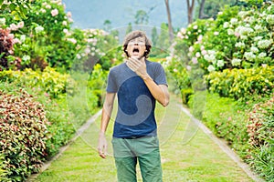 Young man sneeze in the park against the background of a flowering tree. Allergy to pollen concept