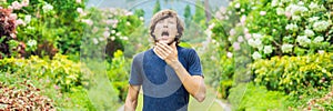 Young man sneeze in the park against the background of a flowering tree. Allergy to pollen concept BANNER long format photo