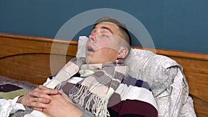 A young man sneezes. The guy is sick lying on the bed with a scarf around his neck