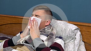 A young man sneezes. The guy is sick lying on the bed with a scarf around his neck