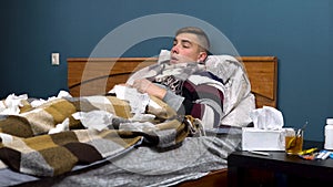 A young man sneezes. The guy is sick lying on the bed with a scarf around his neck. Paper scarves are scattered around