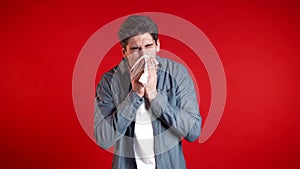 Young man sneezes into tissue. Isolated guy on red studio background is sick