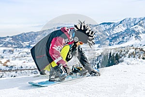 Young man snowboarder in fanny hat sits in the snow and fastens fastenings on a snowboard before the descent