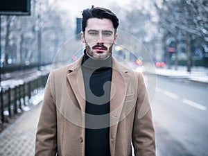 Young man in snowy city, wearing wool coat, looking at camera