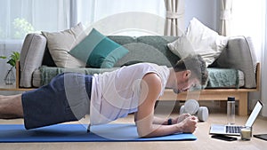 Young man in sports clothes doing plank on blue mat and watching fitness lessons online on laptop. Sport at home