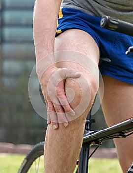 Young man in sportswear on bike in urban park touching his painful knee
